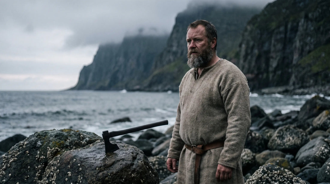 A bearded Norse man in an undyed wool tunic on a rocky shore at dawn, an iron axe resting against the rock beside him