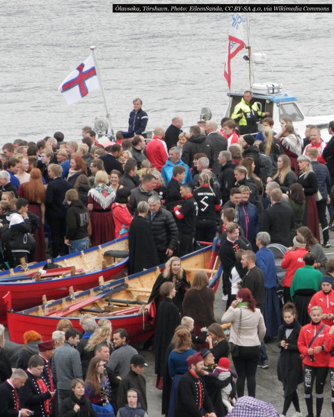 A crowd, many in Faroese national dress, gathered round red wooden rowing boats on the harbour slipway at Ólavsøka in Tórshavn, with the Faroese flag flying from a boat behind (photograph)
