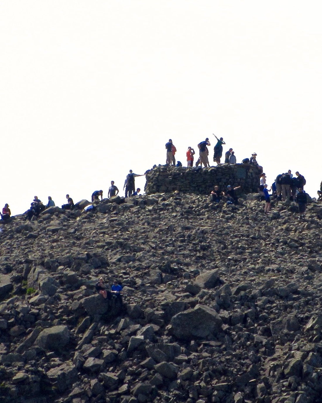 Walkers crowding the rocky summit of Scafell Pike in the Lake District, around the stone summit cairn (photo: Peer Lawther, CC BY 2.0)