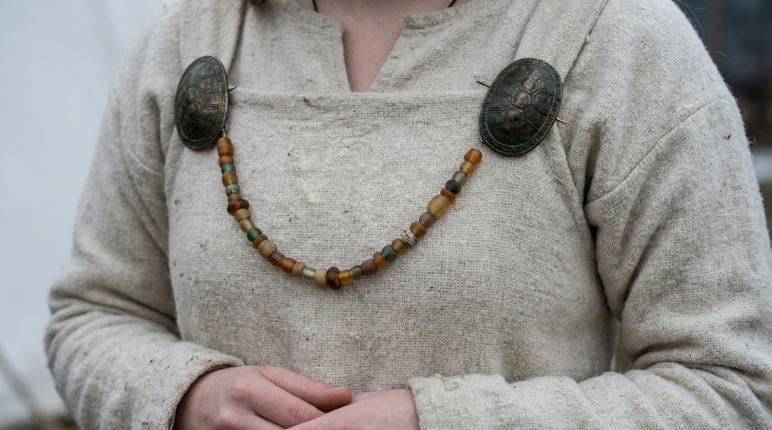 A pair of bronze oval brooches worn at the shoulders of a wool apron dress with a bead string swagged between them