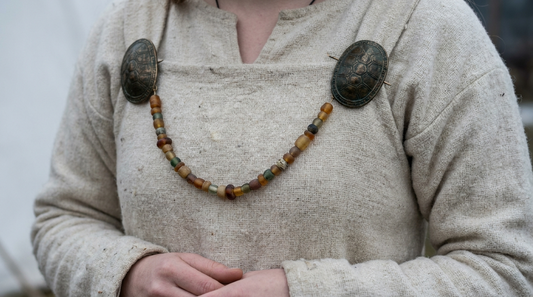 A pair of bronze oval brooches worn at the shoulders of a wool apron dress with a bead string swagged between them