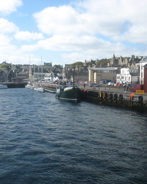 Lerwick harbour under a blue sky with white clouds: a green-hulled boat moored at a quay hung with tyres, yachts beyond, and the town's stone houses rising up the hill behind