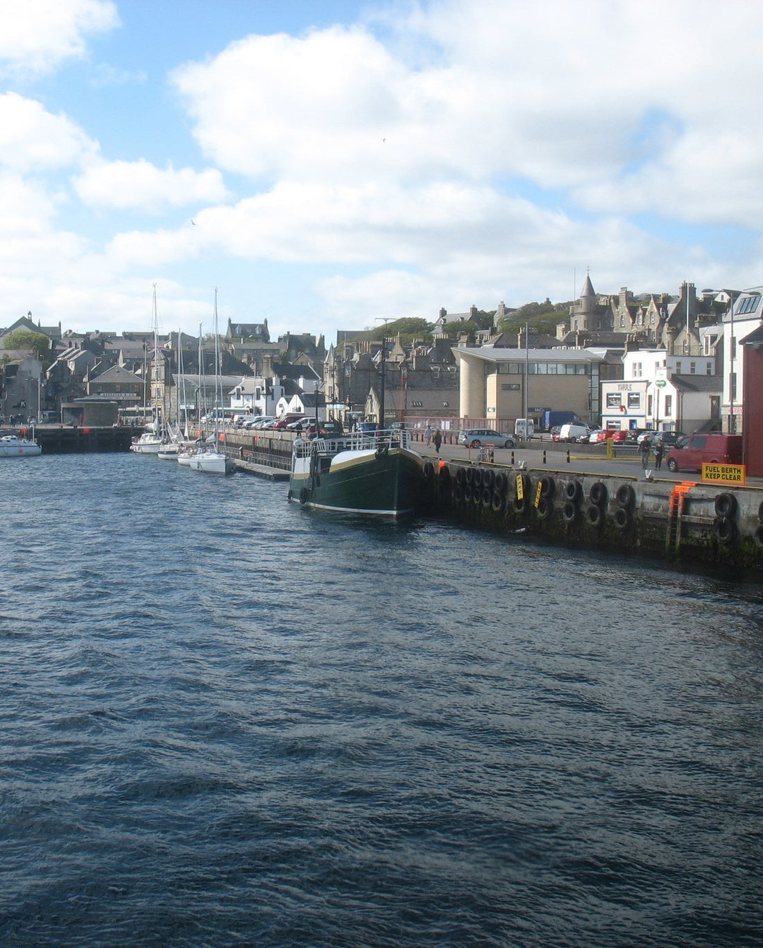 Lerwick harbour under a blue sky with white clouds: a green-hulled boat moored at a quay hung with tyres, yachts beyond, and the town's stone houses rising up the hill behind