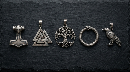 Five silver Norse pendants in a row on slate, a Thor's hammer, a valknut, Yggdrasil, an ouroboros serpent and a raven