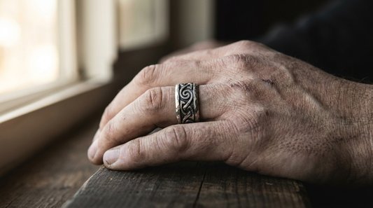 A man's hand on dark oak wearing a wide hand forged silver band with Norse knotwork