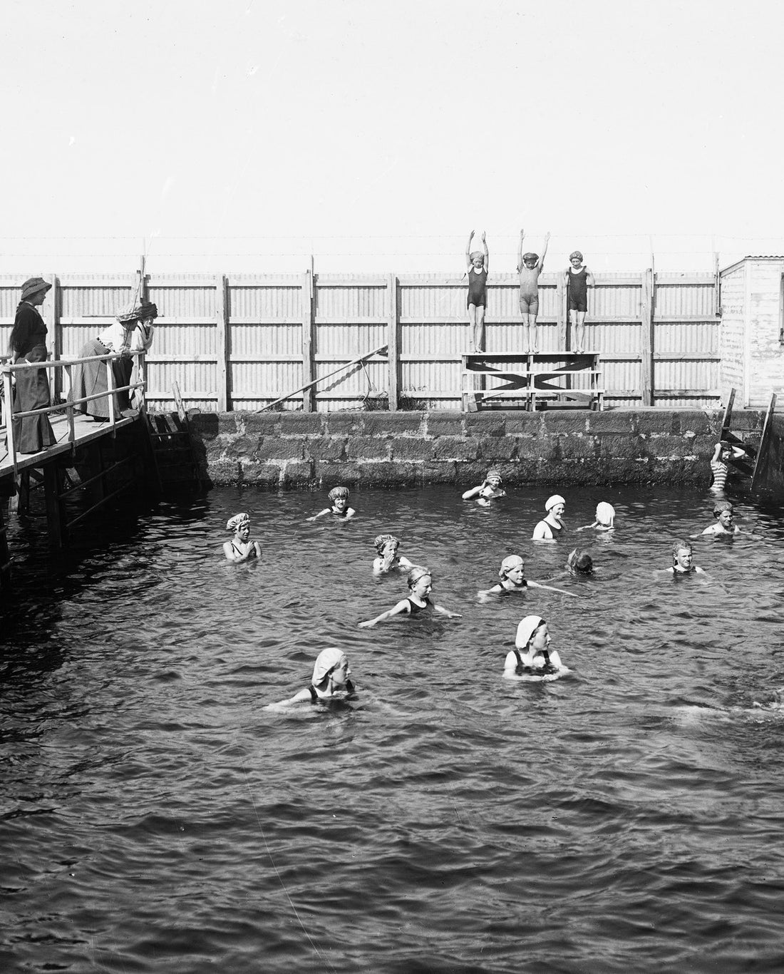 Girls in bathing caps learning to swim in an open-air pool in Laugardalur, Reykjavík, in 1909, three on a platform with arms raised and women watching from a wooden walkway (black-and-white photograph)