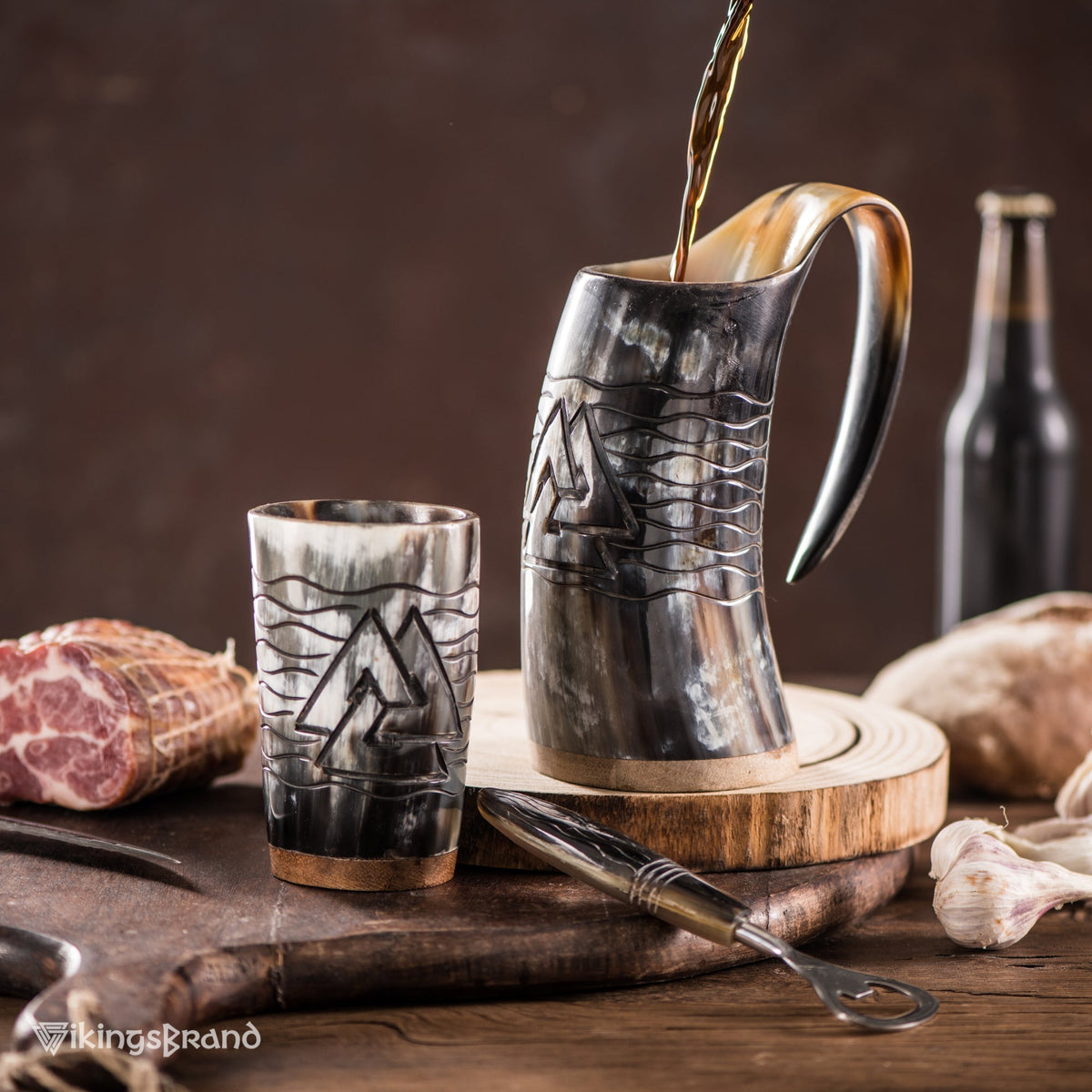 Beer poured into a carved Valknut ox horn mug beside a matching horn shot glass and bottle opener on a wooden board