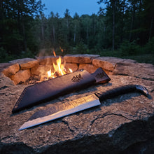 Broken-Back Viking seax knife and its raven-embossed leather sheath lying flat on a stone beside a stone-ringed campfire at dusk