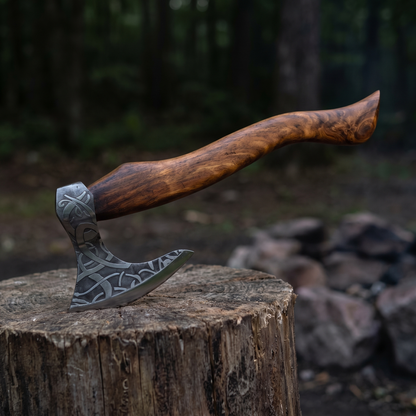 Etched bearded Viking-style axe resting on a tree stump beside a stone fire ring at a forest camp