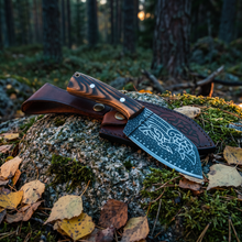 Huginn & Muninn Viking seax knife and its raven-embossed leather sheath on a mossy boulder in a pine forest at dusk