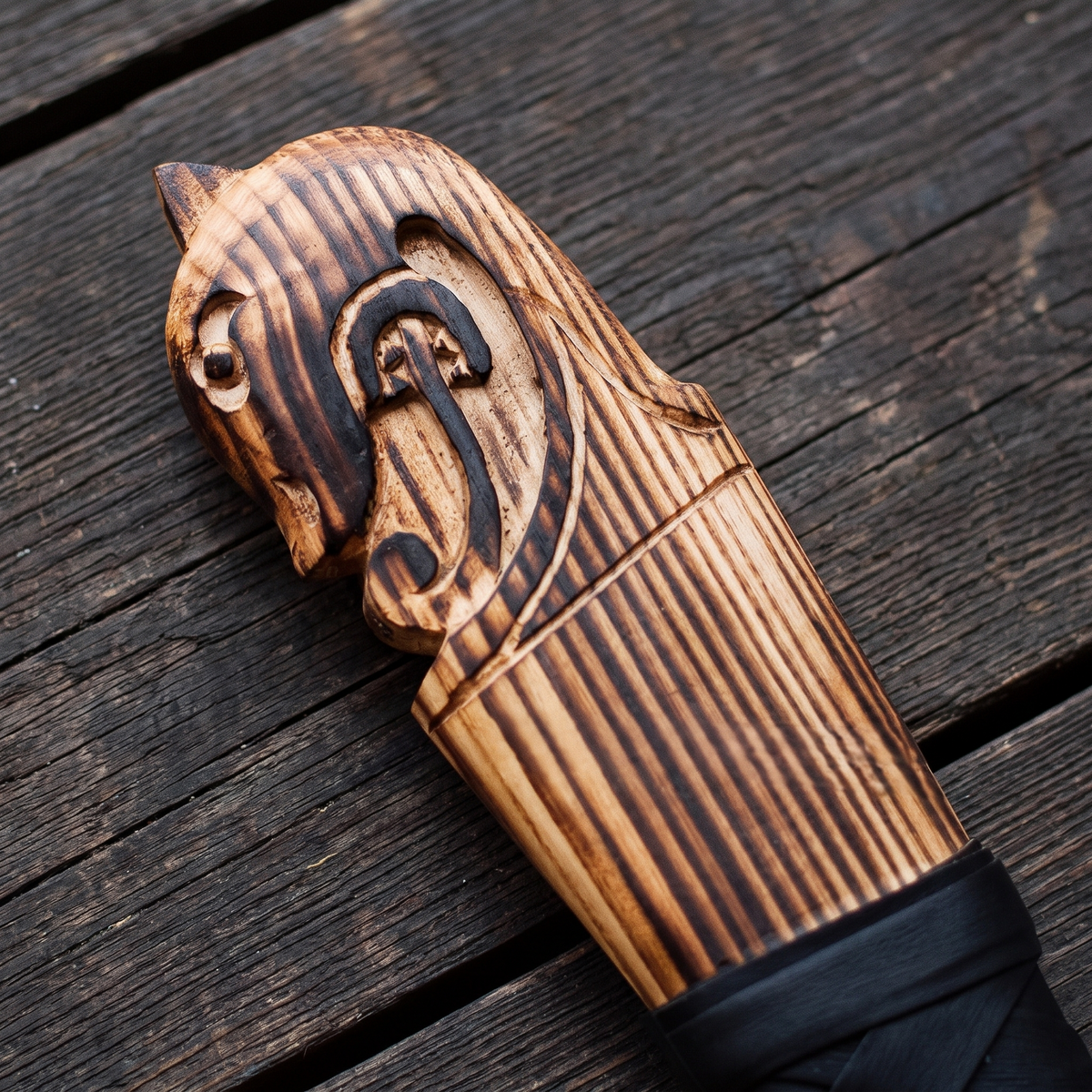 Carved animal-head pommel of the axe handle in scorched light wood above the black wrapped grip