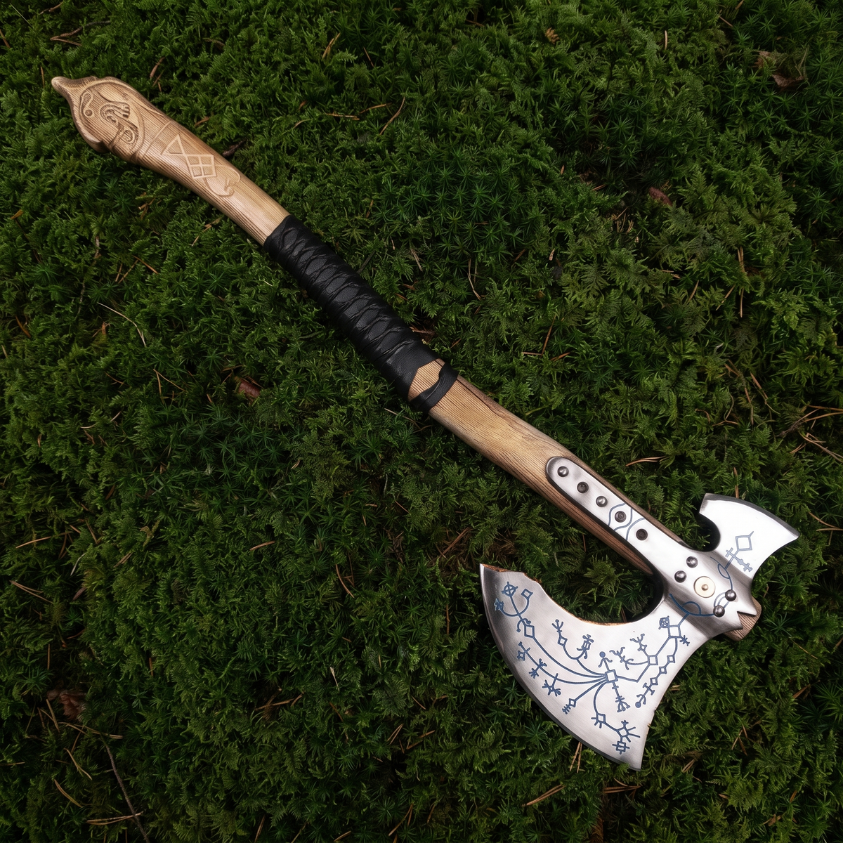 Full-length view of the hand-forged Kratos axe, a Viking-style engraved axe, lying on green moss