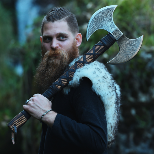 A bearded man in a Viking-style outfit holds a large double-headed axe with engraved silver blades and a dark handle, set against a blurred forest background.