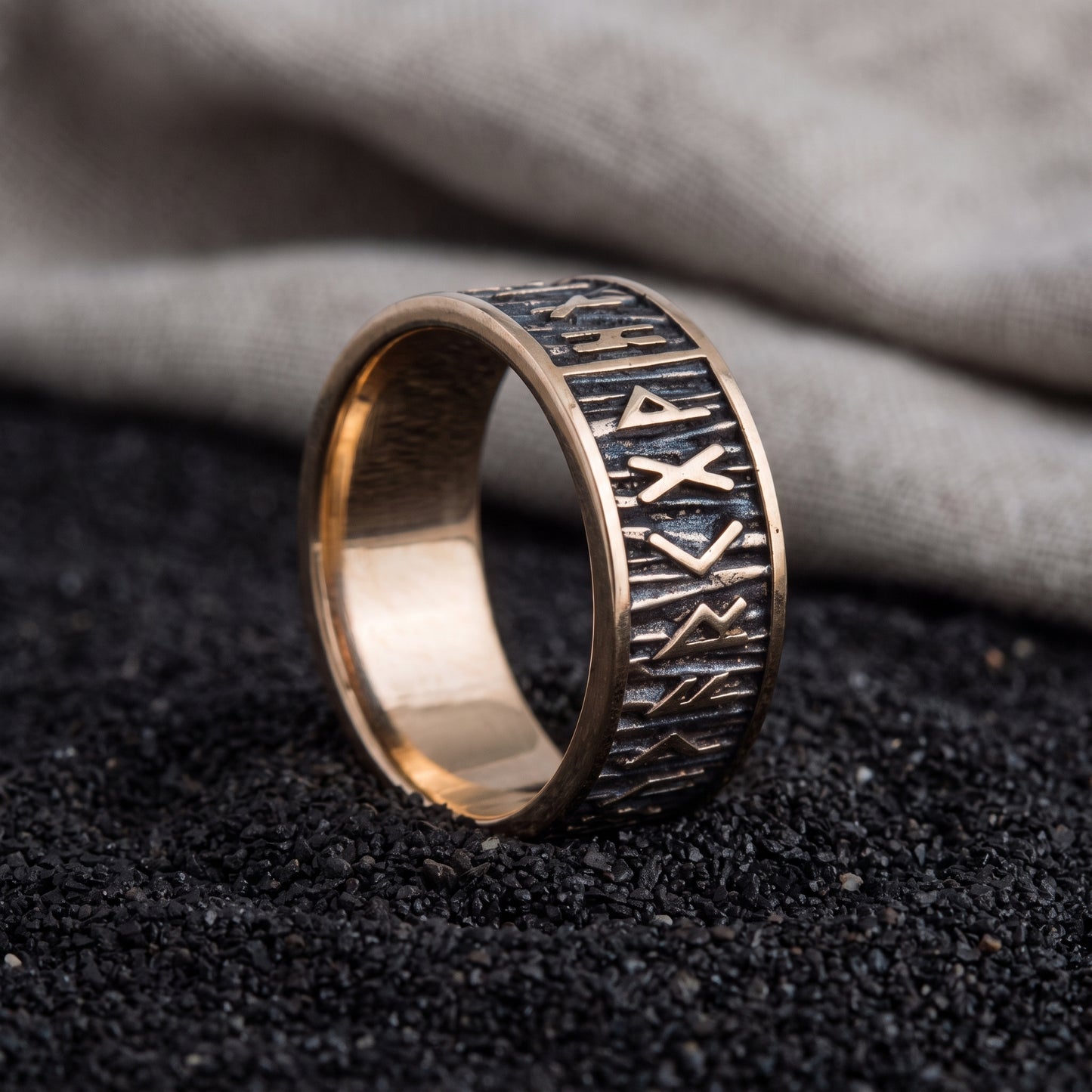 Handmade bronze Elder Futhark rune ring resting on black sand beside undyed grey linen