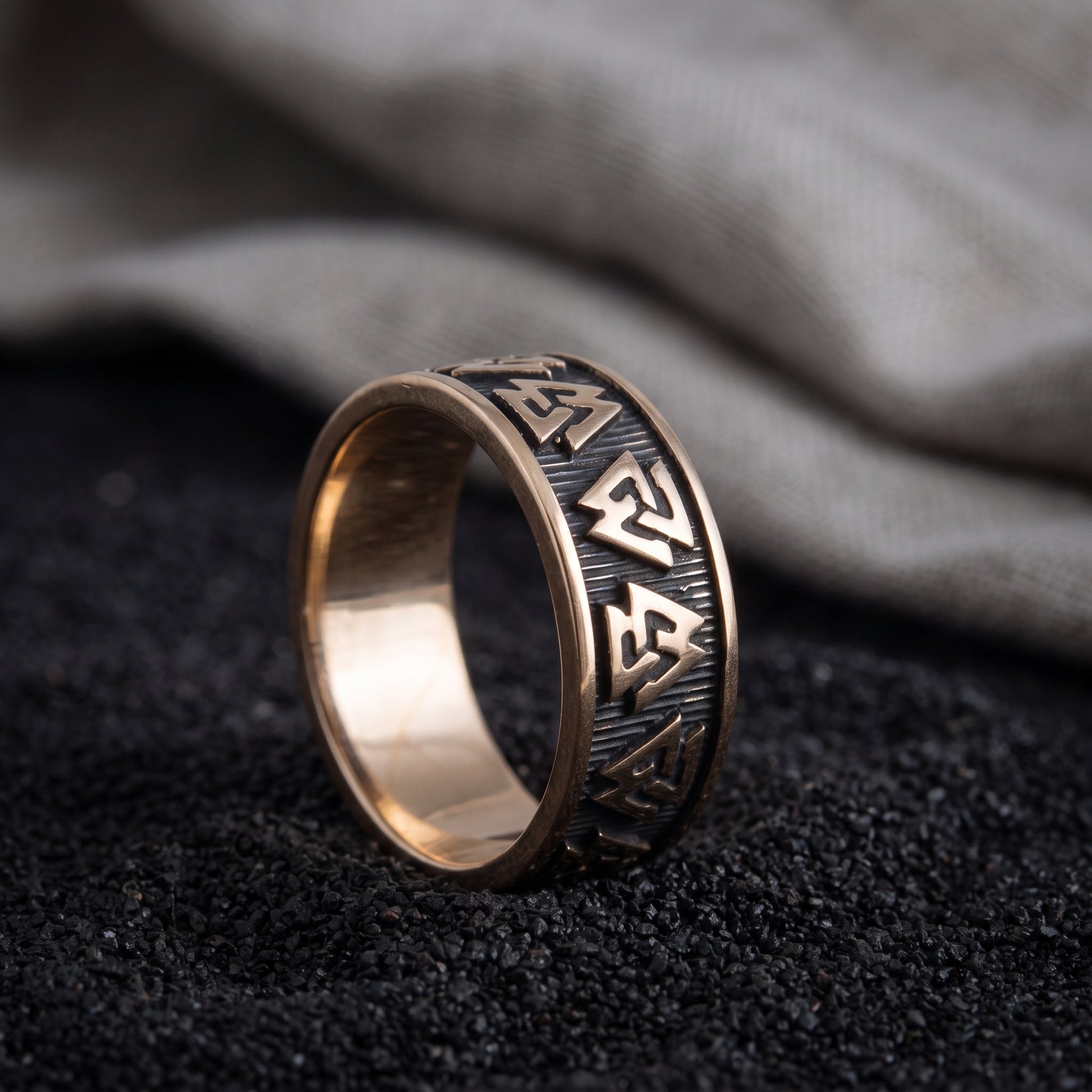 Handmade bronze Valknut Viking ring resting on black sand beside undyed grey linen