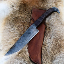 Odin's Raven Viking seax knife with hand-forged carbon steel blade and cast raven's head pommel resting on fur beside its brown leather sheath