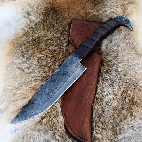 Odin's Raven Viking seax knife with hand-forged carbon steel blade and cast raven's head pommel resting on fur beside its brown leather sheath