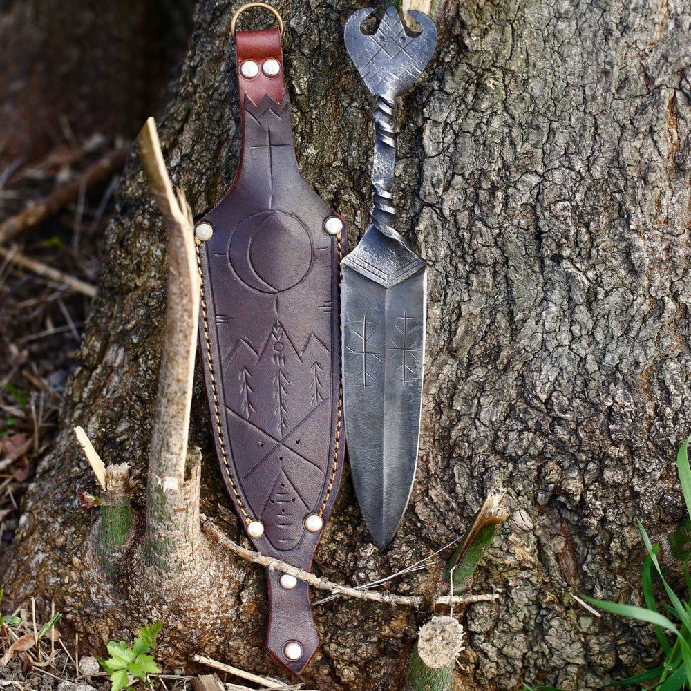Viking dagger and its tooled leather sheath propped against tree bark, showing the crescent and evergreen ornament