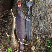 Viking dagger and its tooled leather sheath propped against tree bark, showing the crescent and evergreen ornament
