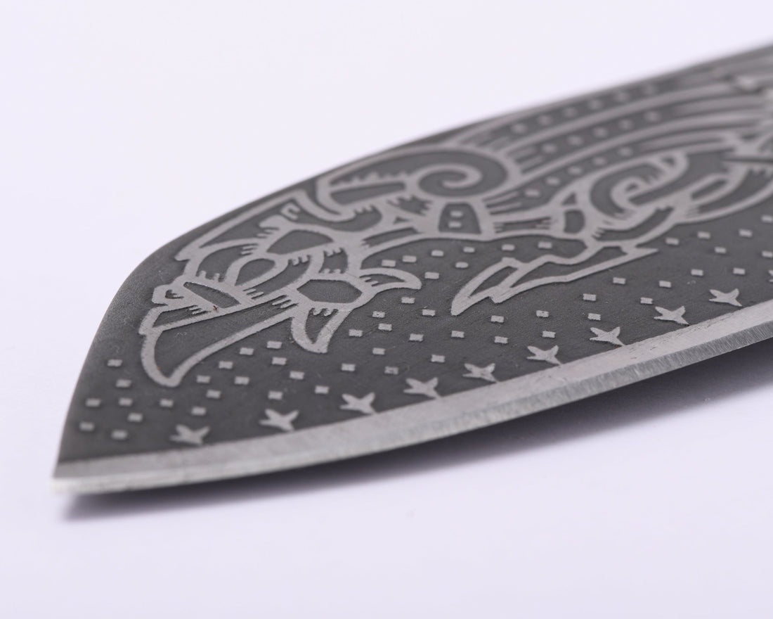 Close-up of Huginn and Muninn etched in interlaced Norse ornament across the darkened carbon steel blade